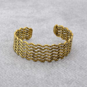 Könnte beinhalten: Ein goldfarbenes Armreif-Armband mit einem wellenförmigen Design. Das Armband hat mehrere Schichten aus wellenförmigen Linien, die ein strukturiertes, offenes Aussehen erzeugen. Das Metall hat eine leicht gealterte, antike Oberfläche.