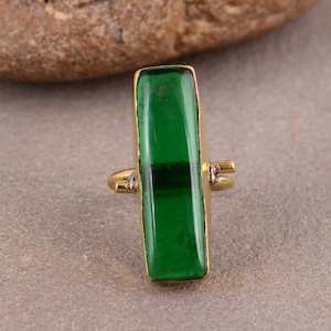 Könnte beinhalten: Ein goldfarbener Ring mit einem rechteckigen, smaragdgrünen Edelstein. Der Ring hat einen langen, rechteckigen grünen Stein, der in einen goldfarbenen Metallrahmen gefasst ist. Das Design ist schlicht und betont das leuchtende Grün des Steins.