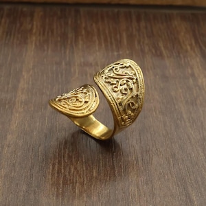 Könnte beinhalten: Ein goldfarbener offener Ring mit zwei ausgestellten, schildförmigen Enden. Jedes Ende ist mit einem komplizierten, erhabenen Rankenmuster verziert. Die Ringschiene ist glatt und poliert und verbindet die beiden strukturierten Abschnitte.