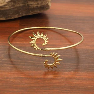 Peut inclure: Un bracelet manchette ouvert de couleur dorée avec deux extrémités en spirale rappelant des rayons de soleil. La bande métallique est fine et circulaire, chaque extrémité s'enroulant vers l'intérieur pour former un motif solaire.