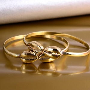 Könnte beinhalten: Goldfarbene Armreifen mit Muscheldesign. Die Armreifen bestehen aus glänzendem, reflektierendem Metall und sind in einer Dreiergruppe angeordnet. Die Muscheln sind mit komplizierten Mustern versehen.