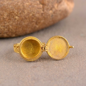 Könnte beinhalten: Ein goldfarbener Medaillonring mit einem offenen, runden Fach. Der Ring hat ein verziertes Band mit Filigranarbeit. Das Medaillon ist geöffnet und gibt das Innere frei. Der Ring liegt auf einer grauen Oberfläche.