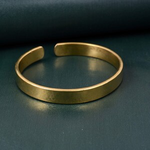 Könnte beinhalten: Ein goldfarbenes Armreif-Armband mit schlichtem, offenem Design. Das Armband hat ein flaches, rechteckiges Profil und eine leicht strukturierte Oberfläche. Die offenen Enden ermöglichen eine einfache Anpassung und das Tragen. Das Armband wird vor einem dunkelgrünen Hintergrund gezeigt.