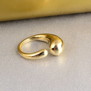 Könnte beinhalten: Ein goldfarbener Ring mit einem einzigartigen tropfenförmigen Design. Der Ring ist an einer Seite offen und hat eine glatte, abgerundete Oberfläche.