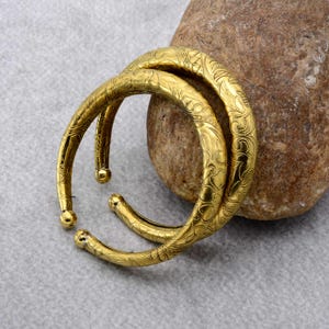 Könnte beinhalten: Zwei goldfarbene Armreifen aus Metall mit einem detaillierten, geprägten Blumen- und Rankenmuster auf der gesamten Oberfläche. Jeder Armreif hat ein abgerundetes, offenes Design mit kleinen, kugelförmigen Endstücken.
