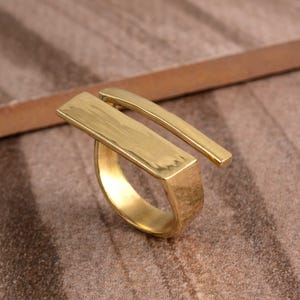 Könnte beinhalten: Ein goldfarbener Ring mit einem einzigartigen Design, bestehend aus einem breiten, strukturierten Band und einer dünnen, geraden Stange, die sich von der Oberseite des Bandes erstreckt.