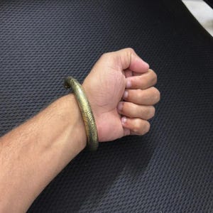 Könnte beinhalten: Ein goldfarbenes Armreif-Armband mit einem aufwendigen, geätzten Design. Das Armband wird am Handgelenk getragen und zeigt seine detaillierten Muster und seinen metallischen Glanz. Die Oberfläche des Armreifs reflektiert das Licht und hebt die gravierten Details hervor.