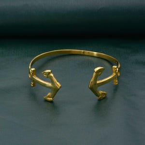 Könnte beinhalten: Ein goldfarbenes Armreif-Armband mit offenem Design. Das Armband hat zwei ankerförmige Enden, jedes mit detaillierten Designelementen. Das Armband besteht aus einem glänzenden, metallischen Material und ist vor einem dunkelgrünen Hintergrund platziert.