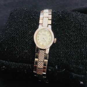 May include: A gold-toned wristwatch with a white face and black Roman numeral markings. The watch has a gold-toned metal band.