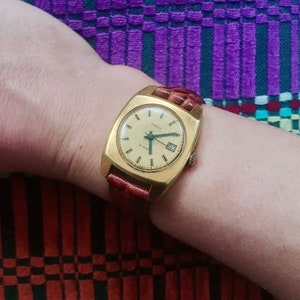 May include: A gold-toned wristwatch with a square face and a brown leather band. The watch face has black Roman numeral hour markers and a date window at the 3 o'clock position. The watch is on a person's wrist.