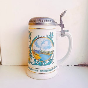May include: White ceramic beer stein with a pewter lid and handle. The stein features a blue and green landscape scene with a river and trees. The text "ПОВОЛЖЬЕ - 1977" is printed on the bottom of the stein.