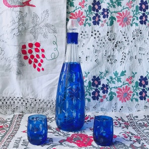May include: A blue glass decanter with a white floral design and two matching blue glass shot glasses. The decanter and glasses are sitting on a white tablecloth with a floral pattern.