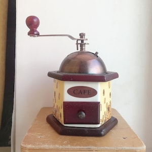 May include: A vintage wooden coffee grinder with a hand crank, a brown wooden base, and a yellow and white ceramic body with a floral design. The grinder has a drawer with a brown wooden front and a metal knob. The word "CAFE" is printed on the front of the drawer.