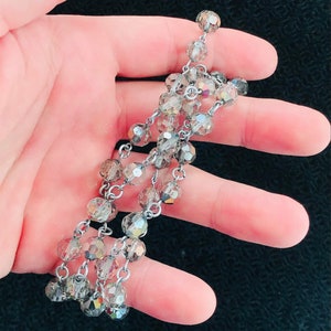 Puede incluir: Un collar de cadena de plata con múltiples hileras de cuentas de cristal facetadas. Las cuentas son transparentes con un ligero brillo iridiscente.