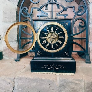 Op de afbeelding: Een zwarte antieke klok met een goudomrand gezicht en Romeinse cijfers. De klok heeft een open, ronde, goudkleurige deur. De klok staat op een stenen oppervlak, met een decoratieve smeedijzeren boog op de achtergrond.