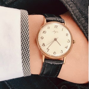 May include: A gold-tone wristwatch with a black leather band. The watch face is white with black Roman numeral hour markers and black hands. The watch face also has the word "Avia" printed on it.