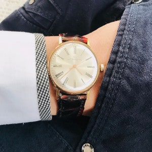 May include: A gold wristwatch with a white face and black Roman numeral hour markers. The watch has a brown leather band with a crocodile pattern.