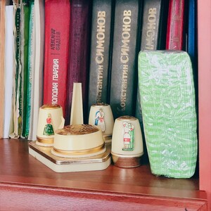 May include: A shelf displays a collection of books with various colored spines, including red, burgundy, and dark green. In front, a decorative set of cream-colored thimbles with gold accents and painted figures is arranged on a rectangular base.