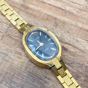 May include: A vintage gold-toned Luch wristwatch with a dark blue oval face. The watch features a gold band with a textured design. The watch face has the brand name "Luch" and the text "15 jewels".