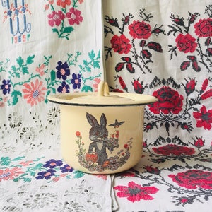 Könnte beinhalten: Ein weißer Emailletopf mit einem schwarzen Hasen- und Vogelmotiv. Der Topf hat einen Deckel und ist mit Blumen und einem roten Apfel verziert. Der Topf steht auf einer weißen Tischdecke mit gestickten Blumen.