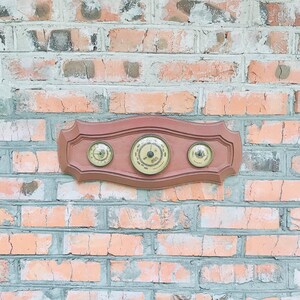 Puede incluir: Una estación meteorológica de pared de madera con tres indicadores analógicos redondos. Los indicadores tienen caras blancas con marcas negras y agujas negras. La estación está montada en una pared de ladrillo.