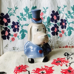 May include: A ceramic dog figurine wearing a blue top hat and jacket. The dog has a cream-colored face and a blue jacket with a red button. The figurine is set against a backdrop of embroidered fabric.