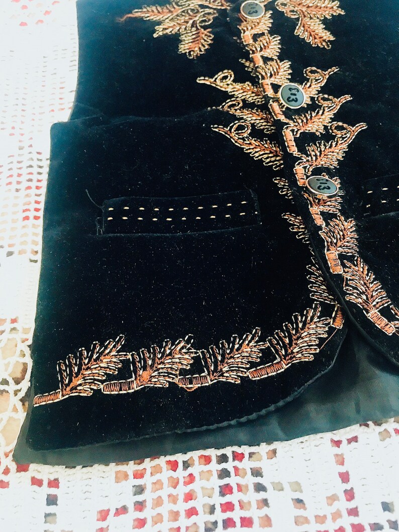 Op de afbeelding: Een zwart fluwelen vest met gouden borduurwerk. Het vest heeft een zak aan de voorkant en drie knopen.