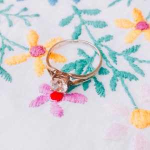 Può includere: Un anello d'argento con una gemma trasparente e rotonda è posizionato su un tessuto bianco ricamato. Il tessuto presenta ricami floreali colorati in giallo, rosa, blu e verde. La fascia dell'anello ha un design decorativo.