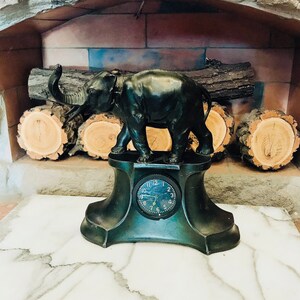 Può includere: Un orologio a forma di elefante nero con un quadrante rotondo e lancette nere. L'orologio è su una base nera con un design decorativo. L'orologio è posto su una superficie di marmo bianco.