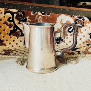 May include: A silver jug with a dark brown handle. The jug has a rounded body and a spout. It is sitting on a white surface.