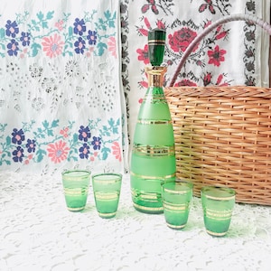 May include: A set of green glass shot glasses and a matching decanter with gold accents. The decanter has a tall, slender shape and a stopper. The glasses are short and round. The set is displayed on a white lace tablecloth.