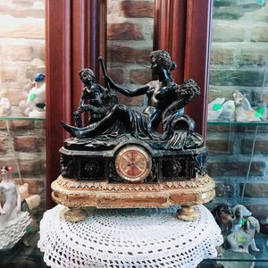 Op de afbeelding: Een zwarte en gouden klok met een sculptuur van drie figuren. De klok heeft een wijzerplaat met Romeinse cijfers en een gouden rand. De sculptuur toont een vrouw en twee kinderen.