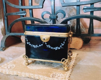 May include: A small, blue glass box with gold trim and a keyhole. The box is decorated with a white floral design and sits on a gold stand with three legs. The box is on a lace doily.