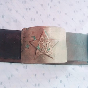 May include: A vintage belt with a large, rectangular buckle. The buckle features a five-pointed star with a hammer and sickle emblem. The belt is made of dark brown leather.