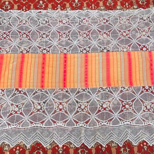 May include: A striped fabric runner with pink, orange, and yellow stripes. The runner is laying on a white crocheted tablecloth.