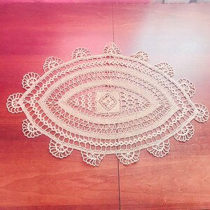 May include: A delicate, off-white crocheted doily with a circular design, featuring intricate lacework and a central oval shape. The doily is laid flat on a wooden surface.