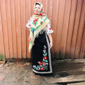 Op de afbeelding: Een vrouw in een traditioneel Oekraïens volkskostuum. Ze draagt een zwarte rok met bloemenborduurwerk, een wit bloesje met borduurwerk en een gele hoofddoek met bloemenpatronen.