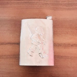 May include: A silver metal flask with a raised design of a person holding a staff. The flask has a screw-on cap.