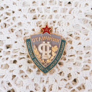 May include: A gold-colored metal badge with a red star and the text "ОТЛИЧНИК СОВЕТСКОЙ ПОТРЕБИТЕЛЬСКОЙ КООПЕРАЦИИ" in Cyrillic script. The badge is on a white crocheted surface.