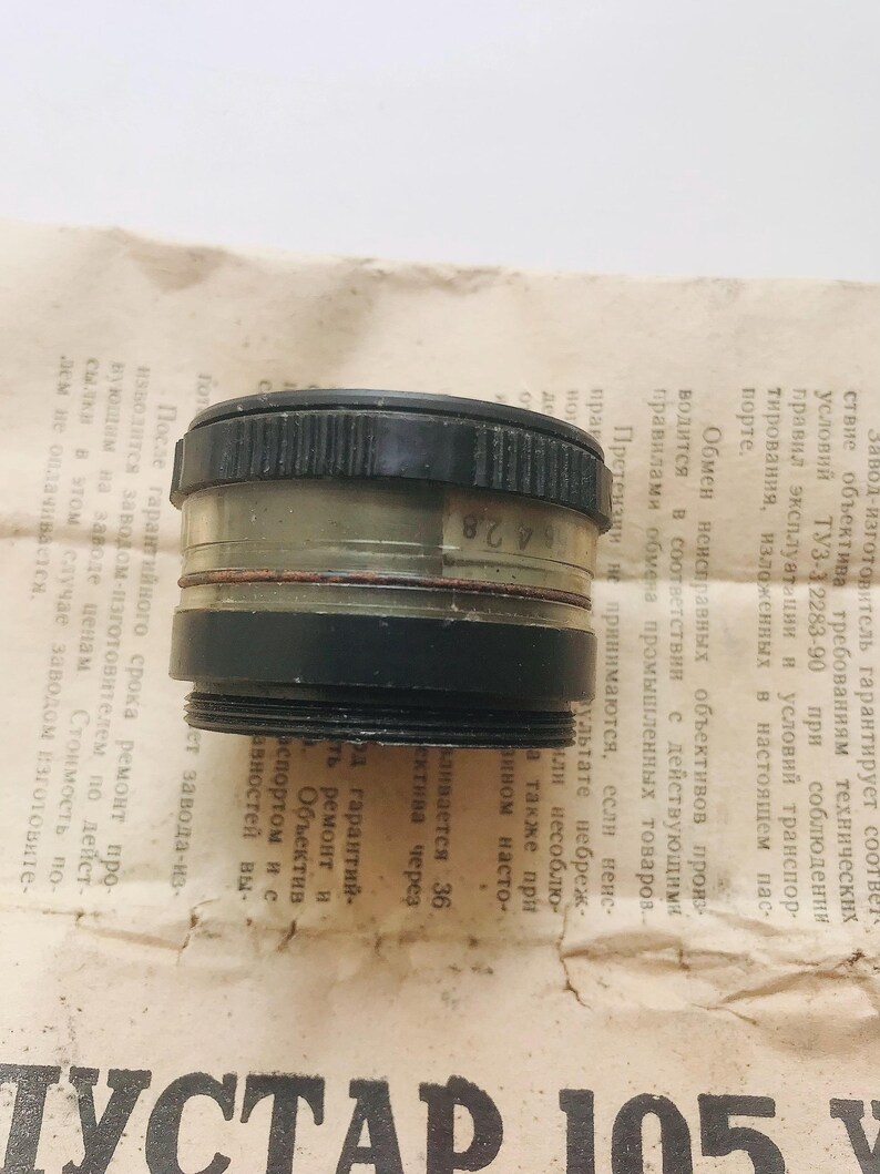 K&ouml;nnte beinhalten: Ein schwarzes und transparentes Kameraobjektiv mit der Nummer "64 2.8" auf der Seite. Das Objektiv ist ein Vintage-Kamerateil.