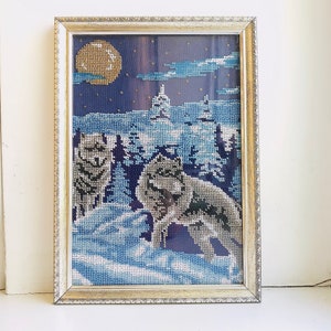 May include: Framed needlepoint artwork depicting a snowy scene with three wolves howling at the moon. The moon is a bright yellow and the sky is a dark blue with white clouds. The wolves are white and gray and are standing on a snowy ground.