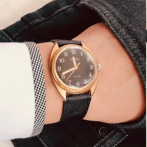 May include: A vintage gold-tone wristwatch with a black face and black leather band. The watch has gold hands and hour markers. The watch is on a wrist, with a white shirt and a black and white checkered cuff visible.