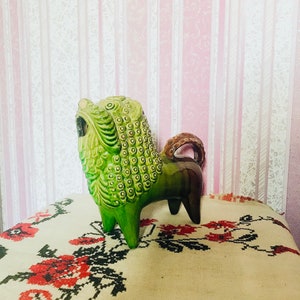 May include: A green ceramic lion figurine with a textured surface and a brown tail. The figurine is standing on a white tablecloth with red and black floral embroidery.