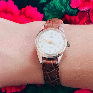 May include: A brown leather wristwatch with a white face and gold-colored hands. The watch has a crocodile-embossed band and a 17 jewels inscription.