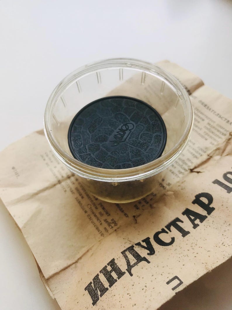 K&ouml;nnte beinhalten: Ein schwarzer Gummideckel mit einer strukturierten Oberfl&auml;che und dem aufgedruckten Wort "INDUSTAR". Der Deckel befindet sich in einem durchsichtigen Glasgef&auml;&szlig;.