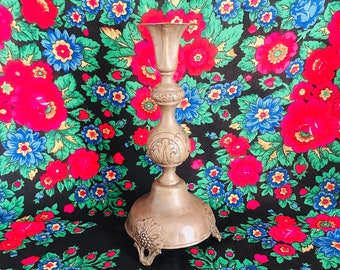 May include: A silver candlestick with an ornate design and three legs. The candlestick is sitting on a black fabric with a floral pattern.