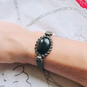 May include: A black oval gemstone bracelet with a silver-toned metal setting. The bracelet has a detailed design with a braided pattern.