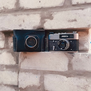 May include: Two vintage cameras, one in a black leather case and the other a silver camera with a black lens and the text "FED 5B" on the top.