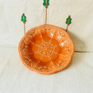Puede incluir: Un plato de cerámica de color terracota con un diseño floral intrincado en relieve. El plato tiene un borde festoneado y está decorado con un patrón central similar a un mandala.