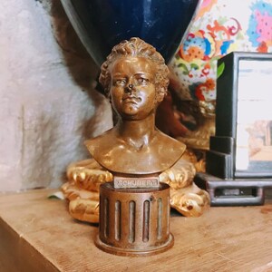 May include: A bronze bust of a young person with the name "SCHUBERT" inscribed on the base. The bust is on a decorative base with vertical grooves.